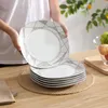 A person is holding a plate from a set of dishes, with more plates stacked on a placemat on a wooden table. Produktbild-Vorschau