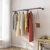 A wall-mounted clothing rack with several garments and a handbag hanging on it, next to a window with sheer curtains and a small wooden table with some fruit. Produktbild-Vorschau