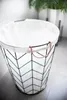 A laundry basket with a white liner and black wire frame with a chevron pattern, featuring copper handles. The basket is placed on a tiled floor next to a potted plant. Produktbild-Vorschau