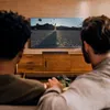 Two people are sitting on a couch watching a TV, with a soundbar on the cabinet below the TV. Anteprima dell’immagine prodotto