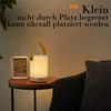 A bedside lamp and a picture frame sit on a small wooden table next to a chair. Produktbild-Vorschau
