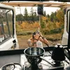 A person is showering outdoors using a portable shower bag attached to a wooden structure, with a van and a natural landscape in the background. Inside the van, cooking equipment is visible. Náhled obrázku produktu