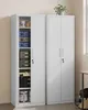 Two tall, narrow white metal cabinets stand side-by-side in a room with a neutral wall and floor. The left cabinet is open, revealing shelves filled with various items like storage boxes, canisters, and tools. The right cabinet is closed. A potted plant si Náhled obrázku produktu