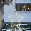 A star-shaped LED light decoration hangs from a tree branch in front of a house with snow on the ground. Produktbild-Vorschau