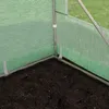 Close-up view of the corner of a greenhouse structure, showing the metal frame, green mesh covering, and dark soil on the ground. Produktbild-Vorschau