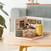 A miniature dollhouse kitchen scene is displayed on a wooden table, with a blurred background of a sofa and a plant. Podgląd zdjęcia produktu
