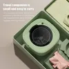 A compact, green washing machine is shown inside a travel case, surrounded by folded clothes and a pink bra. Náhled obrázku produktu