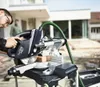 A man is using a miter saw outdoors in front of a modern house. Podgląd zdjęcia produktu
