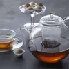 A glass teapot with tea brewing inside, a cup of tea, and chocolates on a dark surface. Produktbild-Vorschau