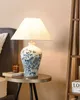 A table lamp with a blue and white porcelain base sits on a small side table next to a bed. A wooden shelf unit is visible on the right. Náhľad obrázku produktu