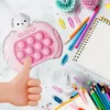 A pink rabbit-shaped portable game with pop-it bubbles, surrounded by stationery items like pencils, paper clips, and a notebook. Produktbild-Vorschau