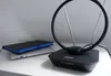 A black active antenna with a circular loop and two extendable rods sits on a white surface next to a closed laptop and a black notebook with a blue spine. Produktbild-Vorschau