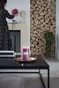 A woman is arranging flowers near a fireplace, with a lit candle on a coffee table in the foreground. Produktbild-Vorschau