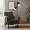 A dark gray armchair with a light gray throw blanket draped over it, next to a black and gold floor lamp with two adjustable heads. The background is a textured gray wall with a framed leaf print above the chair. A window with greenery is visible to the le Náhled obrázku produktu