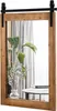 A rectangular mirror with a rustic wooden frame and a sliding barn door hardware is shown. A plant in a vase and some books are placed on a small table in front of the mirror. Náhled obrázku produktu