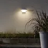 An outdoor wall light illuminates a textured wall, with plants and concrete blocks in the foreground. Produktbild-Vorschau
