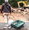 A construction worker in the background, with a lunch box, hard hat, and gloves in the foreground. Podgląd zdjęcia produktu