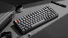 A keyboard is placed on a gray surface with a lens and a pen in the background. Produktbild-Vorschau
