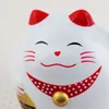 A close-up of a white Maneki-neko (lucky cat) figurine with red ears and a red collar with white polka dots. The cat has a smiling face with closed eyes and a small red heart nose. A gold coin is partially visible in its left paw. Produktbild-Vorschau