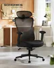 A black ergonomic office chair is shown in a well-lit office setting with a desk and window in the background. Produktbild-Vorschau