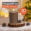 A brown LED candle with a flame sits on a wooden base, surrounded by snow and pinecones, with a blurred Christmas tree and lanterns in the background. Anteprima dell’immagine prodotto