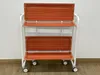 A white metal rolling cart with two orange shelves is shown against a white wall and wooden floor. Anteprima dell’immagine prodotto