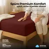 A person sitting on a bed with a burgundy fitted sheet. Produktbild-Vorschau