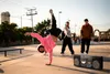 Three young men are in an outdoor skate park setting. One is performing a handstand breakdance move, another is standing behind him, and the third is standing to the right. A large boombox is placed on the ground in the foreground. Produktbild-Vorschau