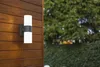 A modern outdoor wall light mounted on a wooden wall, with a blurred background of trees and bushes. Podgląd zdjęcia produktu