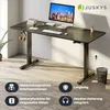 A black height-adjustable desk with a computer, keyboard, mouse, and headphones, set in a home office environment with a plant and a trash can. Produktbild-Vorschau