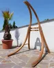 A wooden hammock stand is shown outdoors on a stone patio with a palm tree in a pot to the left. Produktbild-Vorschau