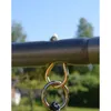 A close-up view of a metal swing set attachment with a bolt and nut on top, and two metal rings and a rope below, set against a blurred outdoor background of trees and sky. Produktbild-Vorschau