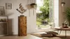 A wooden pedestal with a decorative sculpture on top, placed in a well-lit living room with a large window overlooking a garden. Anteprima dell’immagine prodotto