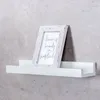 A white picture frame with text is displayed on a white shelf mounted on a gray textured wall. Produktbild-Vorschau