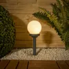 A garden scene with a globe-shaped LED light on a stake, surrounded by white gravel, plants, and wooden planks. Produktbild-Vorschau