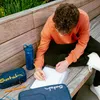 A young person is sitting on a wooden bench outdoors, studying and writing in a notebook. School supplies like a pencil case and a water bottle are laid out next to them. Voir les images du produit
