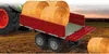 A red trailer filled with hay bales is attached to a green tractor in a field. Produktbild-Vorschau
