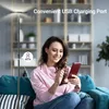 A woman is sitting on a couch and charging her phone using a floor lamp with a USB port. Produktbild-Vorschau