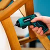 A hand holding a green and black cordless screwdriver, using it to screw into a wooden chair leg. Produktbild-Vorschau