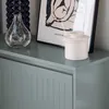 A close-up view of a light gray cabinet with decorative vertical lines on its doors. On top of the cabinet are a clear glass vase, a framed picture, and a white ceramic jar. Náhled obrázku produktu