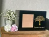 A black urn with a photo frame and a tree design is displayed on a wooden surface, next to a vase of baby's breath and two carnations. Náhled obrázku produktu