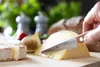 A hand is using a knife to cut a wedge of cheese from a larger block. Other cheeses, a fig, and bread are visible in the background, suggesting a meal or picnic setting. Voir les images du produit
