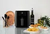 A black air fryer is displayed on a countertop with food items and kitchen utensils. Produktbild-Vorschau