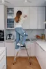 A woman is standing on a stepladder in a modern kitchen, reaching for a cabinet. Náhled obrázku produktu
