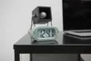 A digital alarm clock is shown on a black table, with a laptop and a decorative object in the background. Podgląd zdjęcia produktu