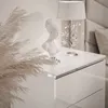 A white bedside table with a bust and a lamp on it, with pampas grass in the foreground. Produktbild-Vorschau