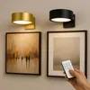 Zwei zylindrische Wandleuchten, eine goldene und eine schwarze, beleuchten gerahmte Kunst an einer hellgrauen Wand, mit einer Hand, die eine Fernbedienung hält Náhled obrázku produktu