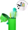 A green dinosaur-shaped pencil case is open, with pens and fluffy decorations inside. A circular inset shows a white pencil holder filled with colorful pens and a panda face. Voir les images du produit