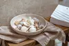 A bowl of seashells rests on a textured cloth on a wooden surface, with a stack of books in the background. Produktbild-Vorschau