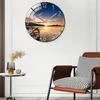 A round wall clock with a sunset scene is displayed on a white wall next to a chair and a small table with a vase. Voir les images du produit